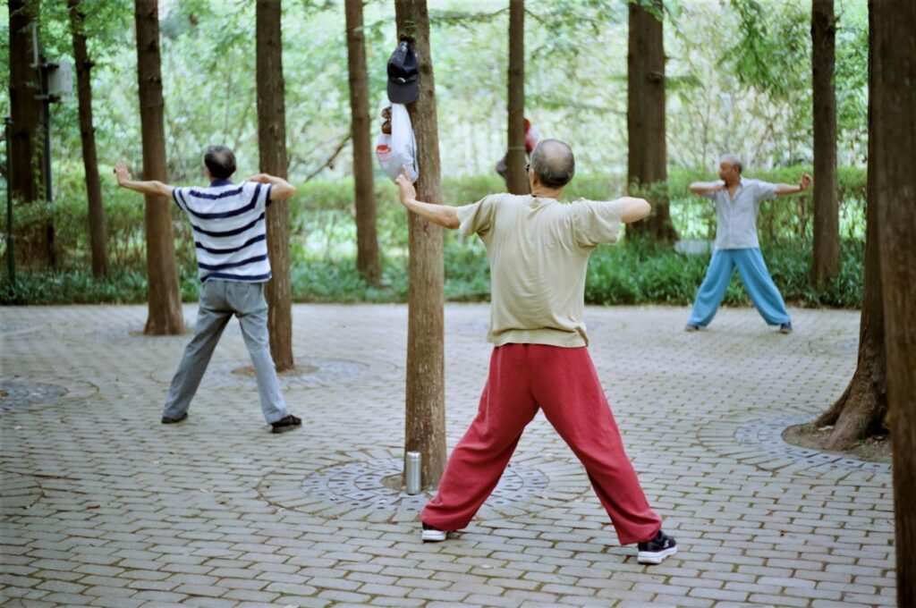 Three Chinese adults performing tai chi exercises outdoor under trees